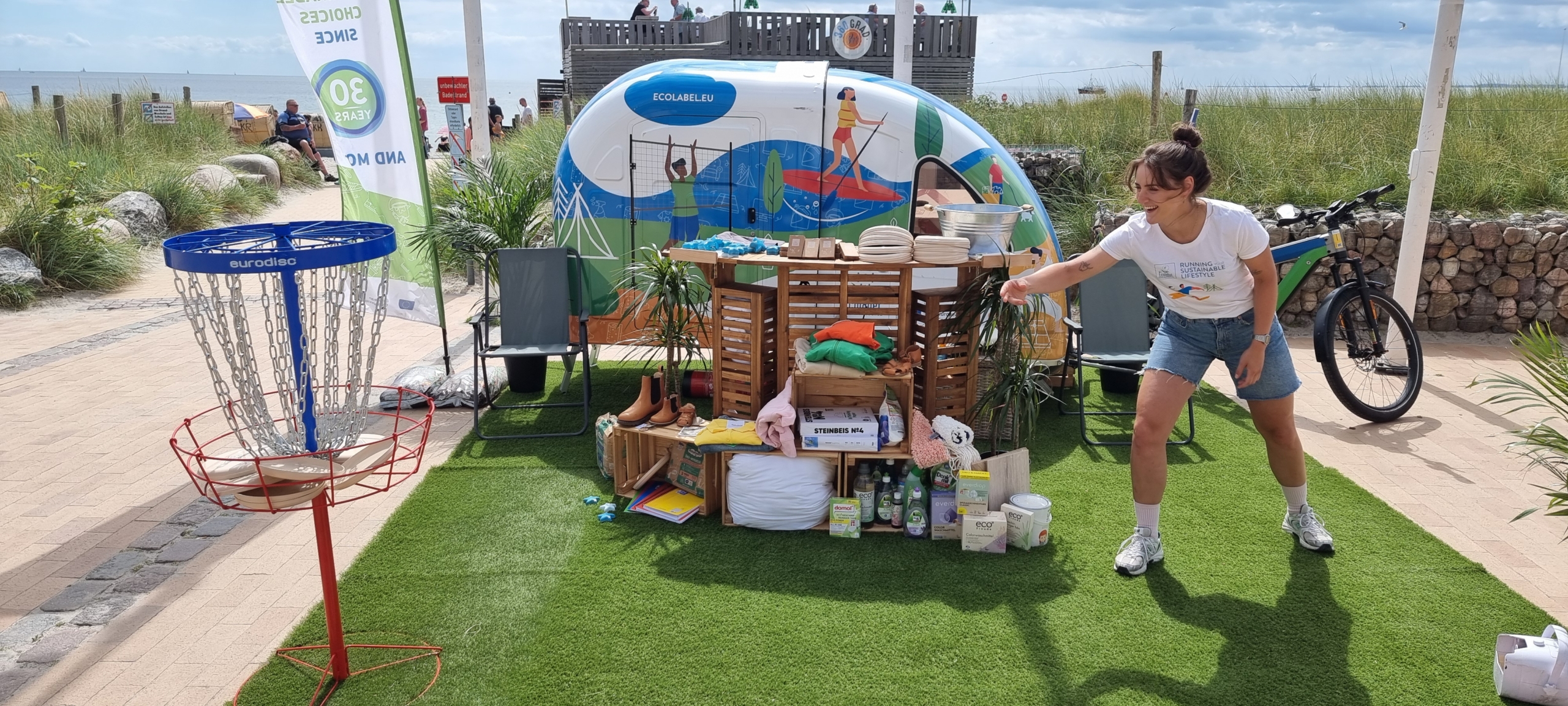 Der EU Ecolabel Infostand am Strand mit zertifizierten Produkten; eine Frau nutzt das dazugehörige Frisbeespiel