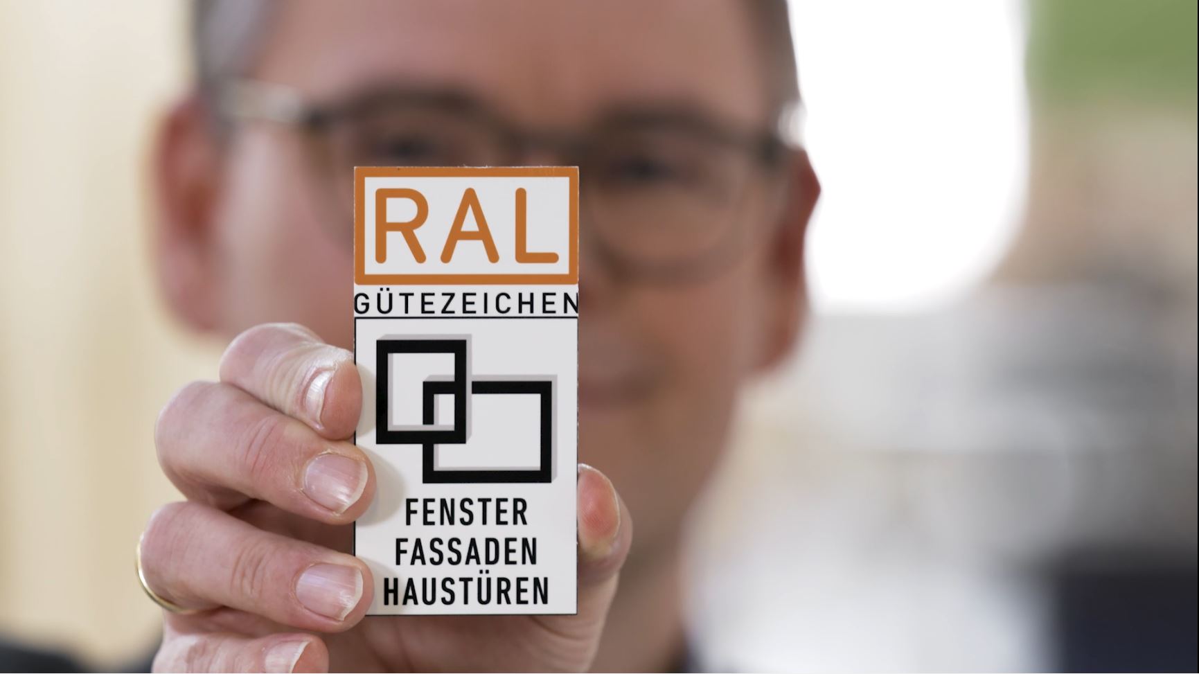 Unscharf zu sehende Person hält das RAL Gütezeichen Fenster, Fassaden, Haustüren hoch